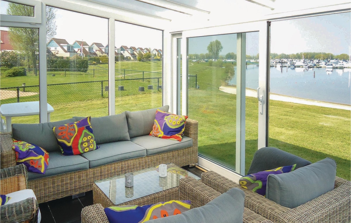 A bright winter garden features large glass windows providing views of the lush green lawn and marina. Comfortable seating is arranged around a glass table, with colorful cushions adding a touch of style.