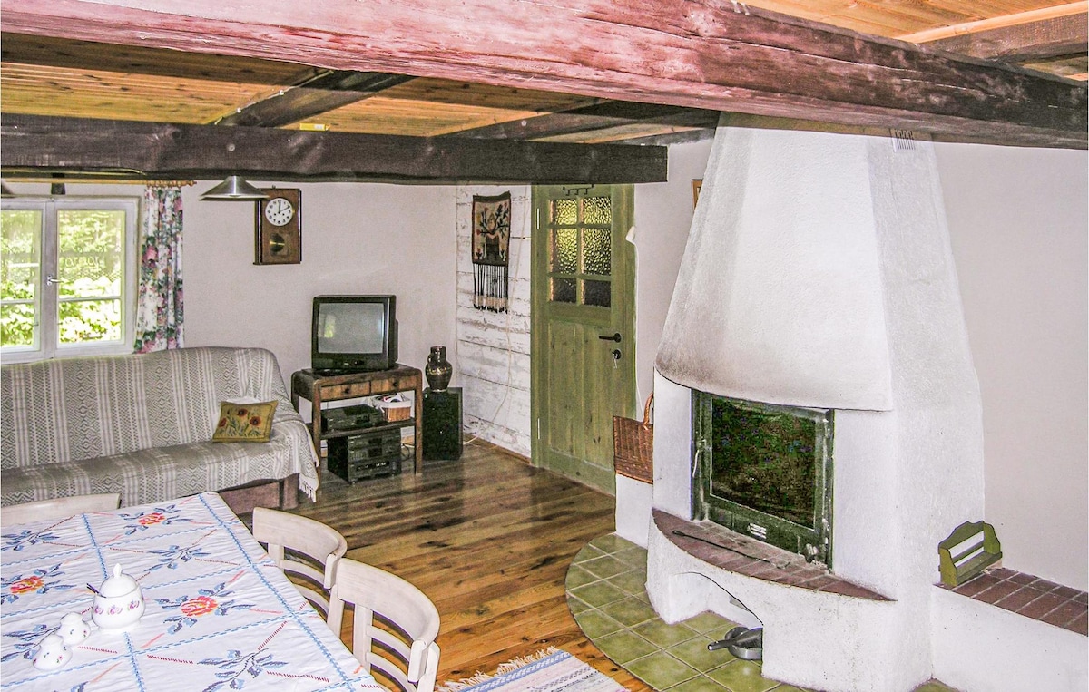 A cozy living area features wooden beams and a rustic fireplace, with a television and a wooden cabinet nearby. A large dining table is adorned with a floral tablecloth, and sunlight filters through the window, illuminating the natural wood flooring.