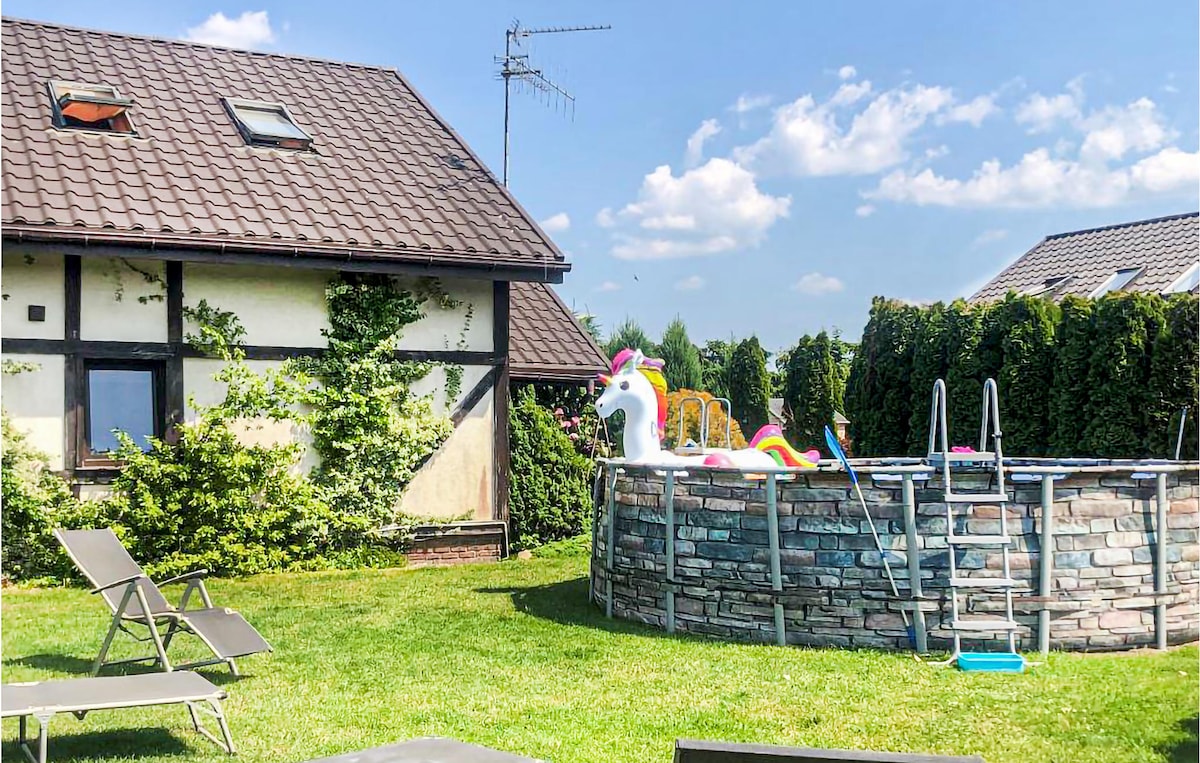 A large above-ground swimming pool is surrounded by a grassy area, featuring colorful inflatable swim toys. Sun loungers are arranged nearby for relaxation. The cottage's exterior is visible, showcasing a charming design with a tiled roof and partially obscured greenery.