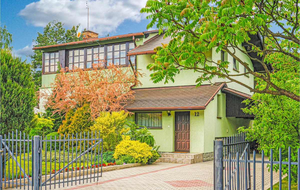 The exterior of a green cottage is framed by lush gardens and a well-maintained fence. A welcoming wooden door leads into the home, while large windows offer glimpses of the interior. Vibrant plants and shrubs accentuate the property, creating a serene outdoor environment.