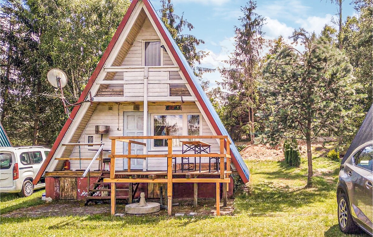 A wooden cottage with a distinctive triangular roof is situated on a grassy plot. A wooden deck with chairs is visible at the entrance, while trees surround the property. A satellite dish is mounted on the side, contributing to the rustic charm.