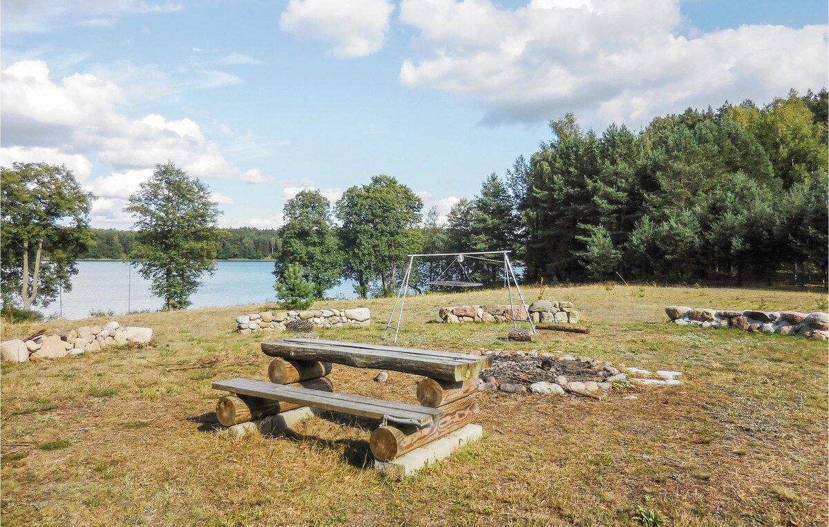 An outdoor area features a wooden picnic table with attached benches and a fire pit made of stone. A swing set is visible nearby, framed by trees and a lake in the background. The landscape is open and lightly grassed.