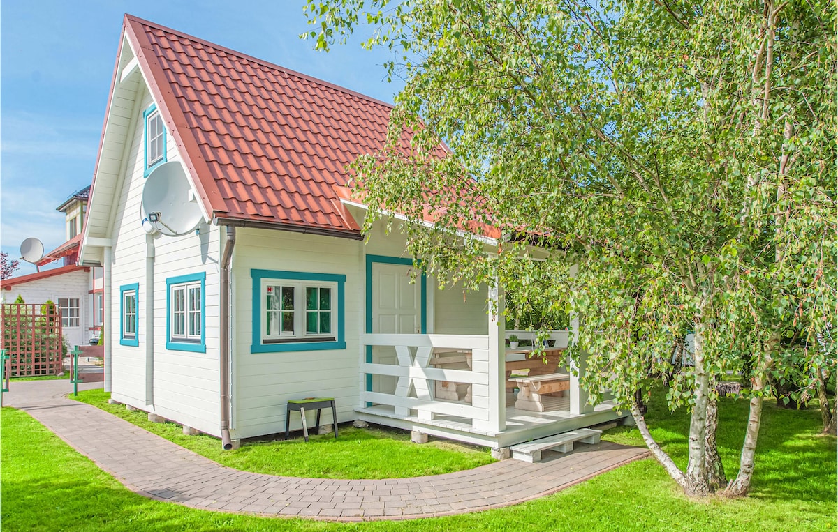 A charming holiday home is presented with a steeply pitched roof and white wooden cladding. A covered porch with seating is visible, surrounded by green grass and trees. Pathways lead to the entrance, highlighting the inviting outdoor space.