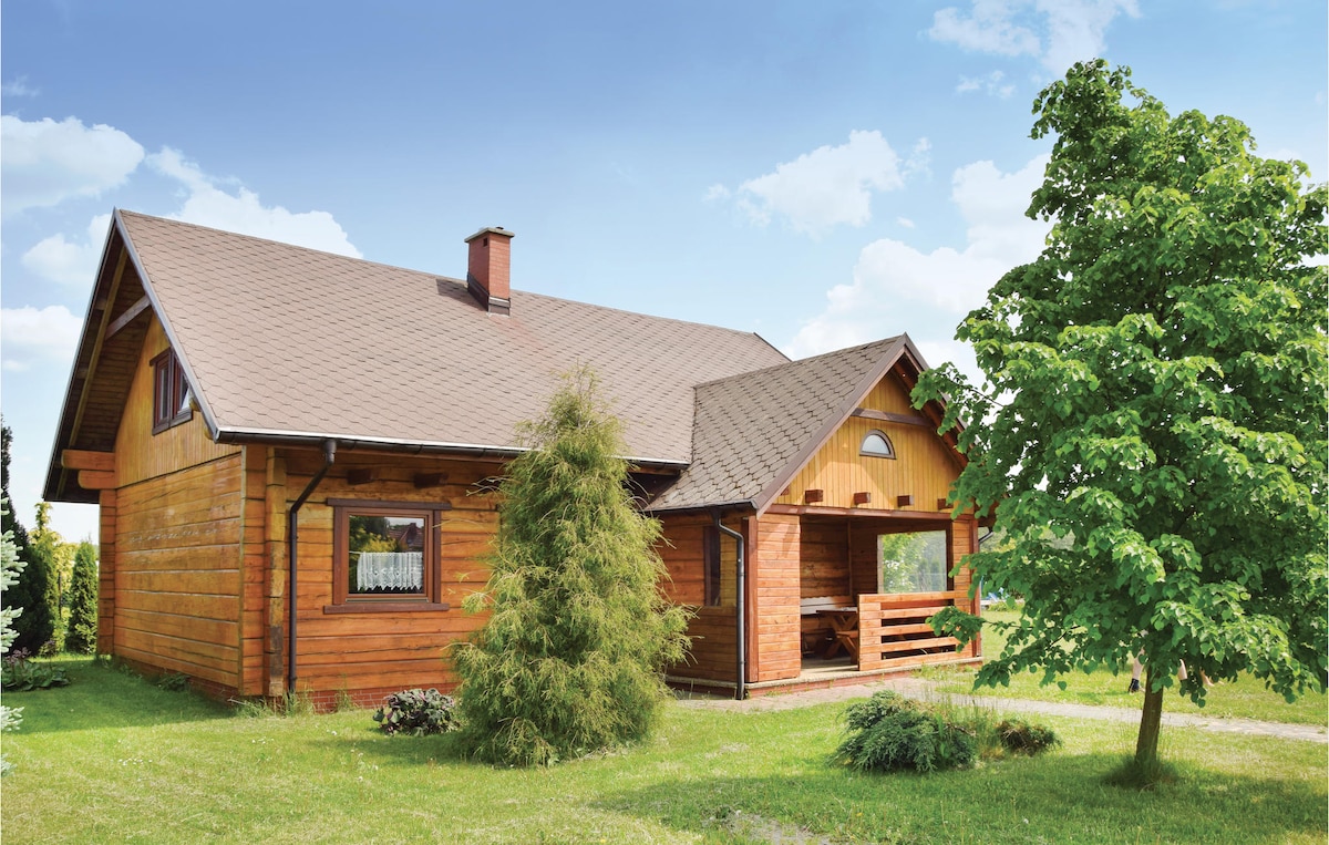 The wooden beam house is set against a bright sky, featuring a sloped roof and large windows. A covered terrace extends from the front, surrounded by lush greenery, including a variety of trees and shrubs that complement the home's natural aesthetic.