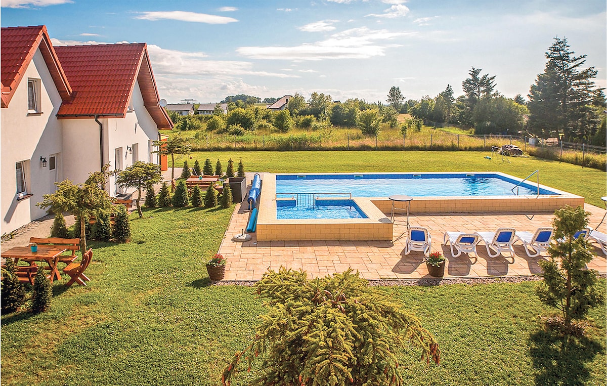 A spacious outdoor area surrounds a large seasonal pool, accentuated by lounge chairs and shaded seating arrangements. Lush green grass and well-maintained landscaping enhance the relaxation space. The pool area is bordered by trees and offers a serene backdrop for various outdoor activities.