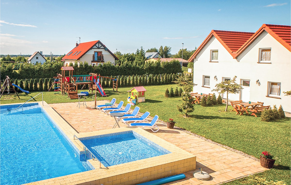 A large outdoor pool is framed by lounge chairs on a sunlit patio. Nearby, a colorful playground structure with swings and slides complements the manicured lawn. Spacious holiday homes are visible in the background, surrounded by greenery and a peaceful atmosphere.