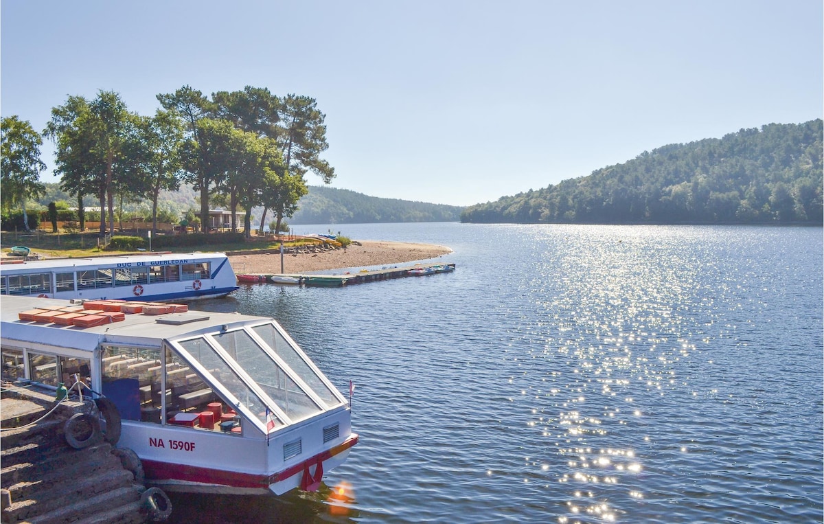 The image depicts a serene lakeside scene at Lac de Guerledan, with a blue sky reflecting on calm waters. A small boat is docked, and sandy beaches are visible in the distance, surrounded by lush green trees and rolling hills.