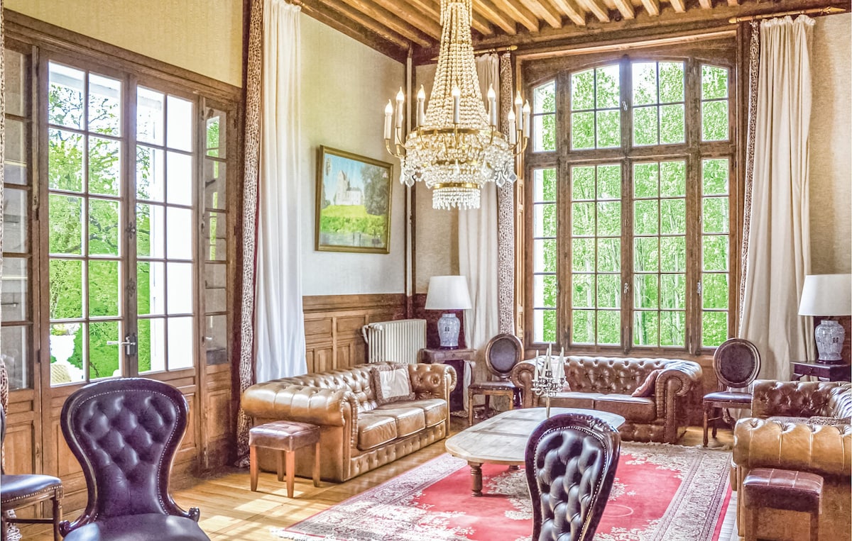 A spacious living room is adorned with elegant brown leather seating and a large chandelier. Tall windows frame views of the lush landscape outside, while an ornate rug anchors the room. Decorative lamps and a painting add character to the refined space.
