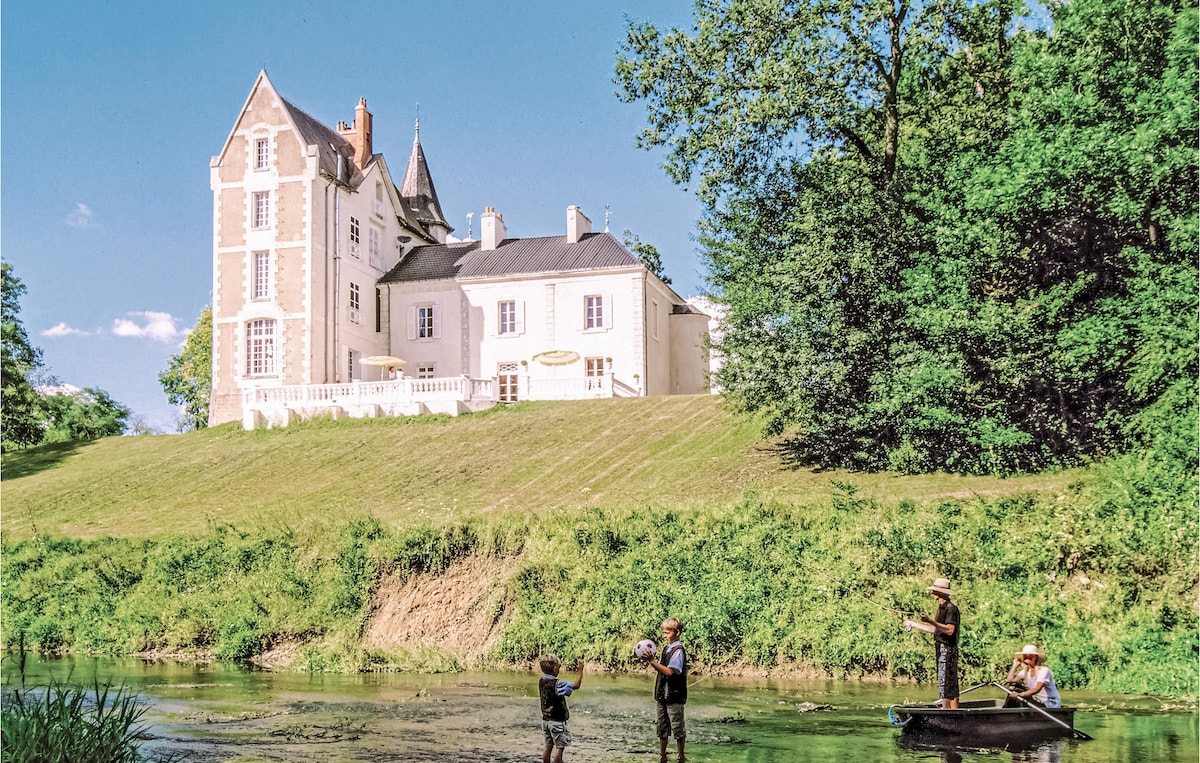 The elegant château is set against a lush green backdrop, near the Indre River. A group of individuals enjoys fishing, with one child wading in the shallow waters while another person paddles a small boat. The architecture features a mix of classic and modern design elements.
