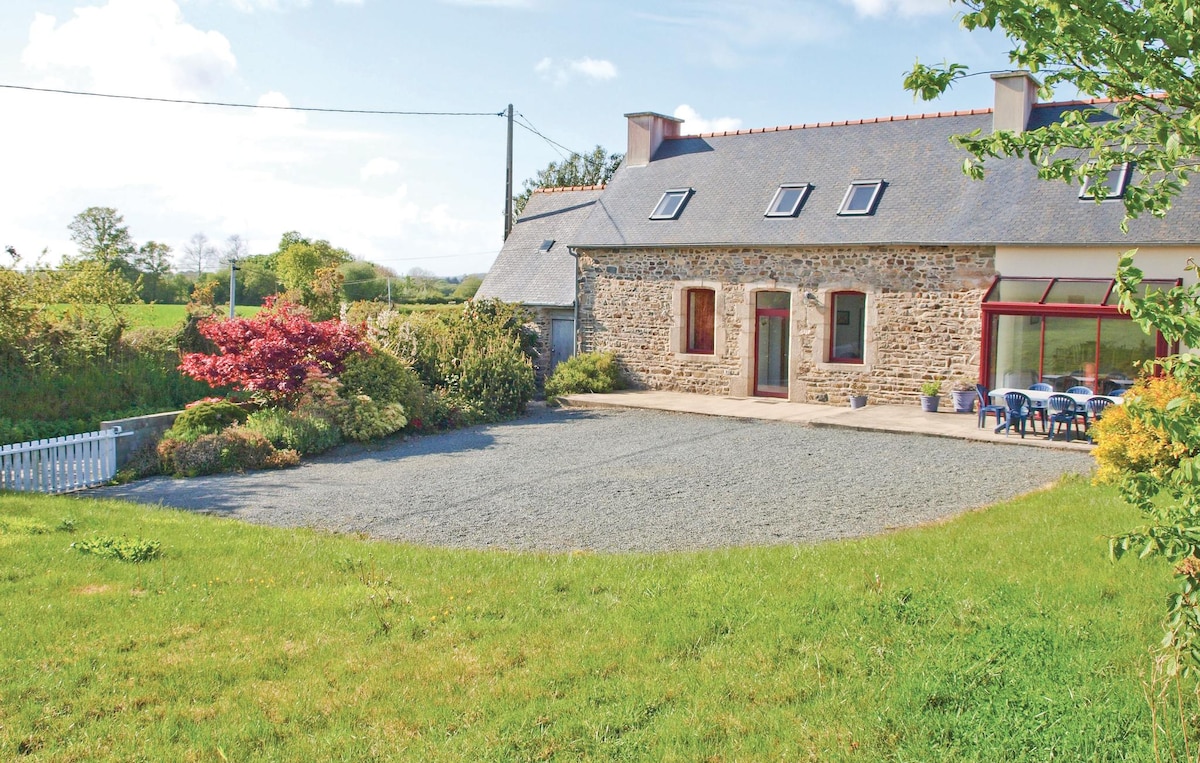 A long stone cottage is set in a rural landscape, featuring a gravel driveway and surrounding green grass. A well-maintained garden with various plants is visible in front, along with a small seating area of blue chairs positioned against the cottage's facade.