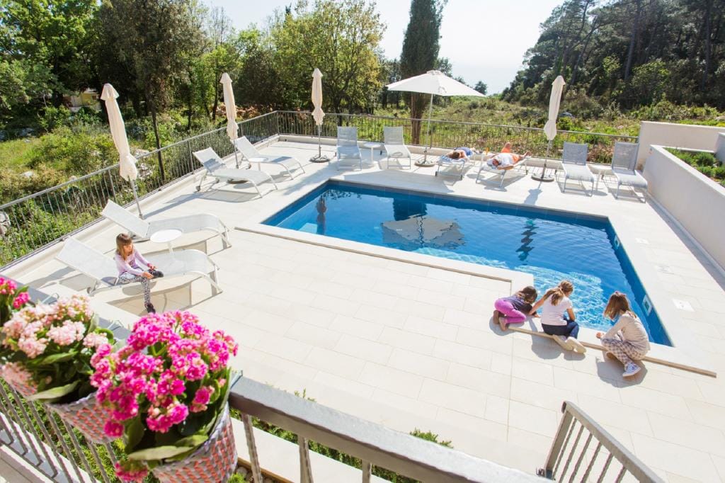 A shared outdoor pool is surrounded by a sun terrace featuring sunbeds and parasols. The area is complemented by vibrant flowers in hanging pots, adding a touch of color. Children play nearby, enjoying the serene atmosphere within the greenery.