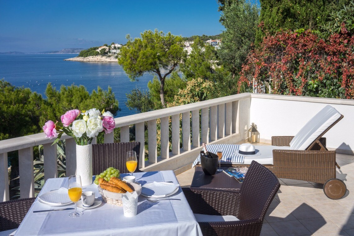 A terrace is presented with a dining table set for breakfast, featuring fresh flowers, pastries, and juice. A comfortable lounge chair is positioned nearby, overlooking the serene sea and coastal landscape, enhanced by greenery and vibrant foliage.