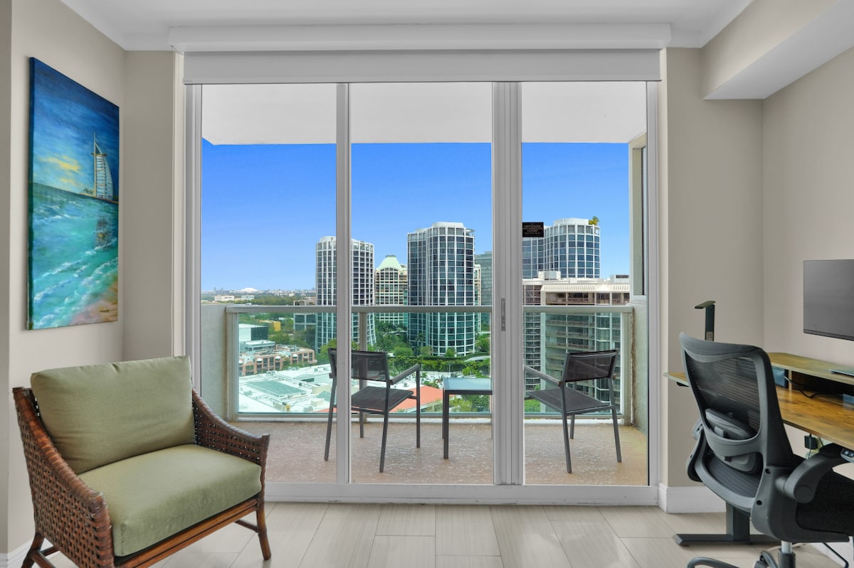 A bright interior space features a comfortable armchair positioned next to a workspace with a desk and office chair. Large glass doors offer a view of the balcony overlooking the cityscape, with outdoor seating visible. Artwork adorns the wall, enhancing the serene atmosphere.