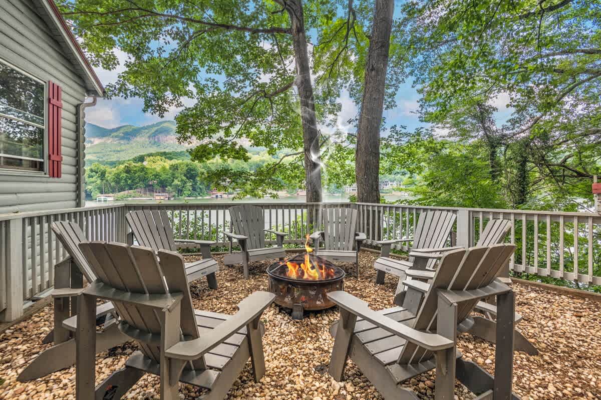 A fire pit surrounded by Adirondack chairs is set on a gravel surface, providing an inviting space for gathering. Lush greenery and trees frame the area, while scenic views of the lake and mountains can be seen in the background.