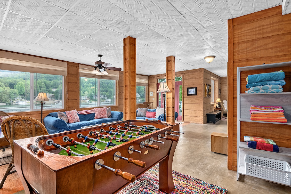 A bright and spacious living area is showcased, featuring a foosball table in the foreground. Large windows allow for ample natural light and provide views of the surrounding greenery and lake. Comfortable seating areas are arranged with inviting blue sofas and side tables.