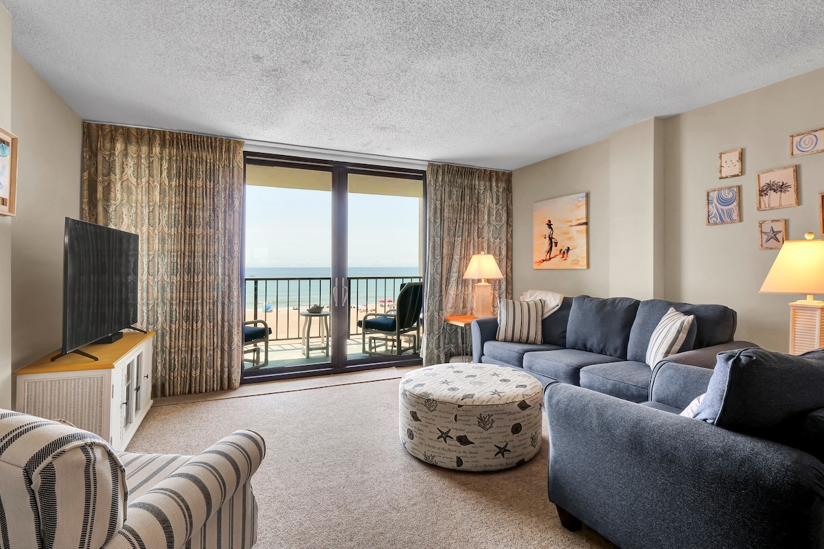 A spacious living area is designed for relaxation, featuring a comfortable dark sectional sofa and a round ottoman with a nautical pattern. Large glass doors open to a balcony with ocean views, while soft natural light illuminates the space through sheer curtains.