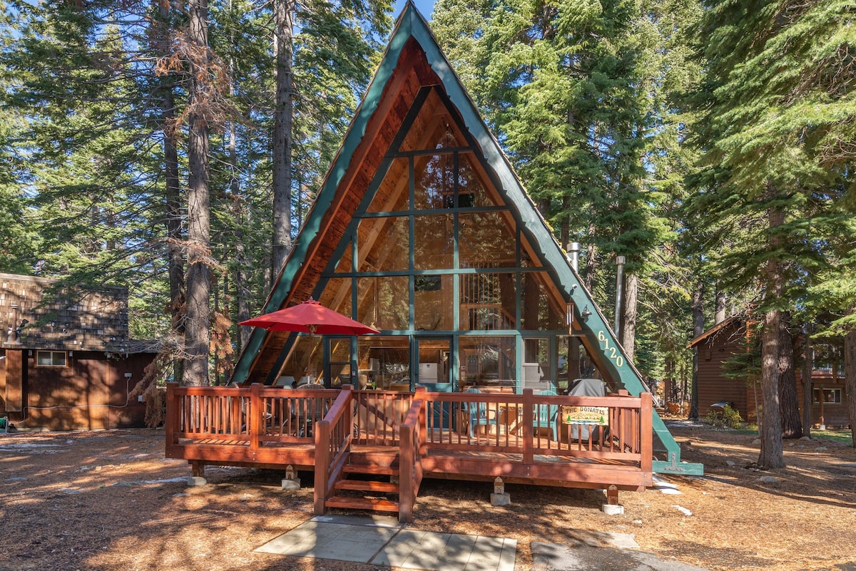 The A-Frame cabin is surrounded by tall trees, featuring a prominent glass front that allows for ample natural light. A spacious wooden deck with a red umbrella provides an outdoor gathering space, while a stone pathway leads up to the entrance.