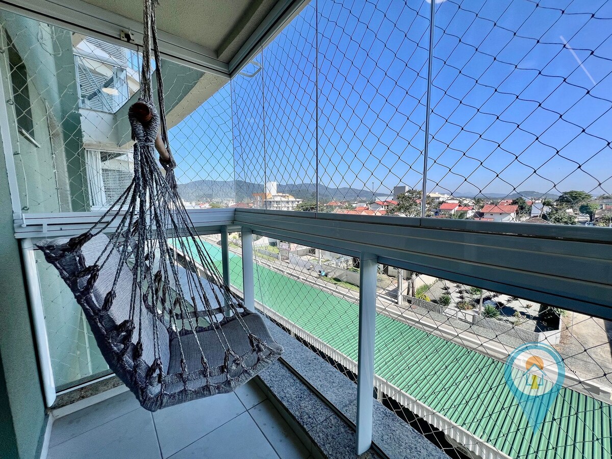 A comfortable balcony with a hammock is visible, featuring a protective netting. The view includes buildings and distant mountains, set against a clear blue sky. Sunlight illuminates the space, enhancing the feeling of openness and relaxation.