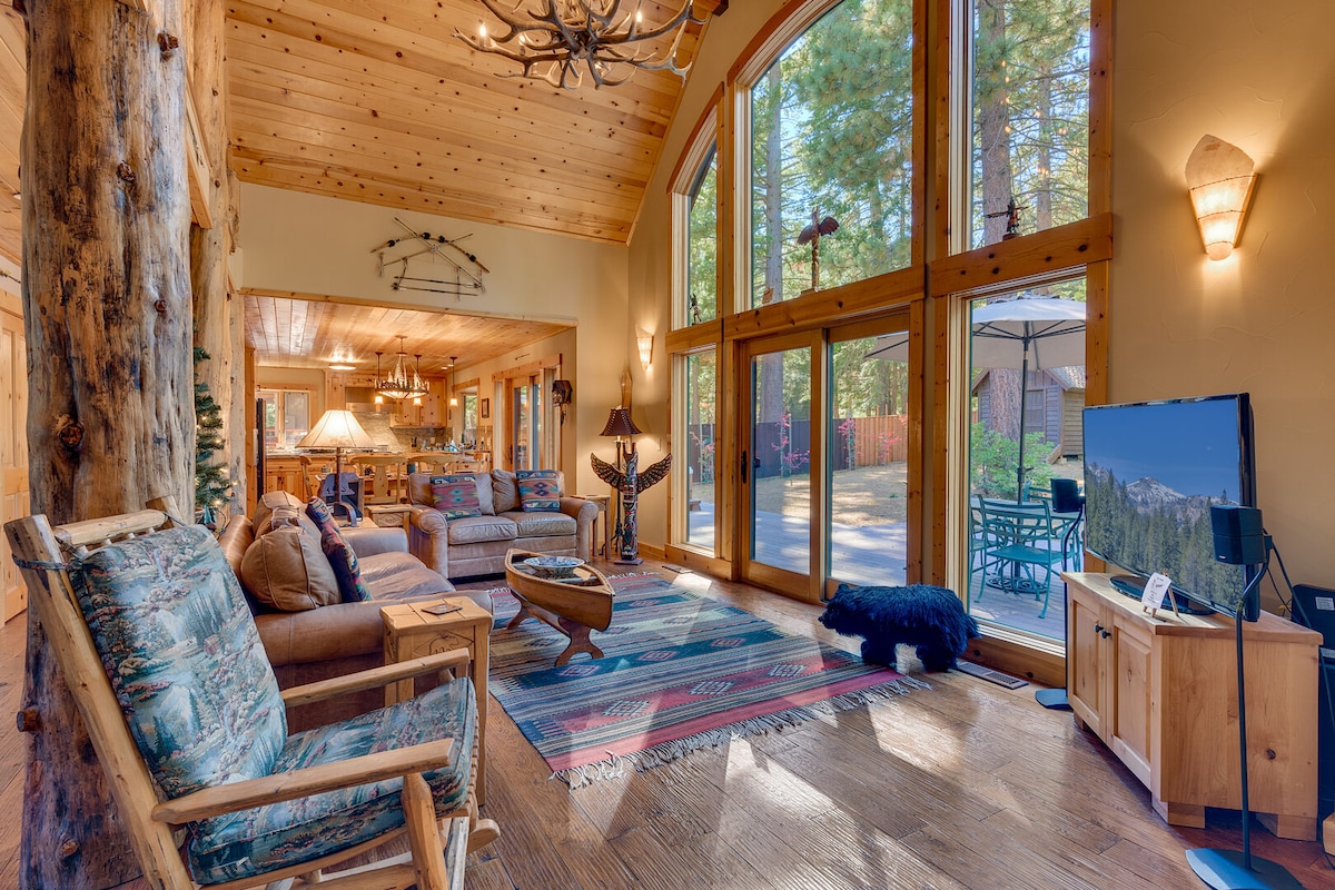 A spacious living area features large glass doors that open to a view of the outdoors. Natural wood elements, comfortable seating, and a decorative area rug contribute to the rustic lodge ambiance. A flat-screen TV is positioned near the seating area.