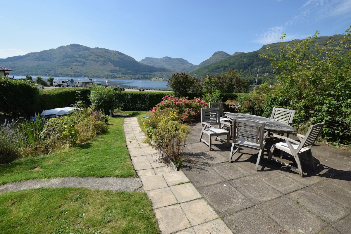 A spacious patio area is surrounded by landscaped gardens, featuring a dining table with multiple chairs. The view overlooks Loch Goil, framed by lush green mountains in the distance. The path leading to the patio is lined with neatly trimmed grass and vibrant flowers.