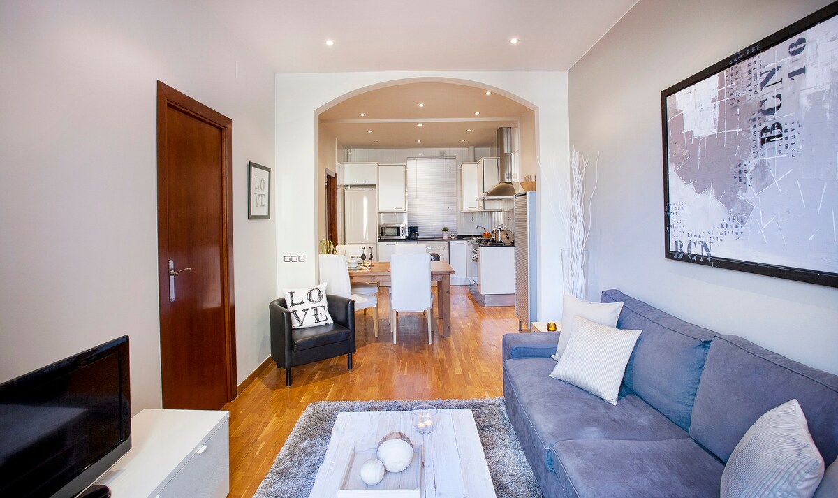 A bright living area showcases a comfortable grey sofa complemented by decorative cushions. A light wooden floor extends through the space, leading to a fully equipped kitchen. A white coffee table featuring decorative items sits in front of the sofa, while soft lighting illuminates the room.