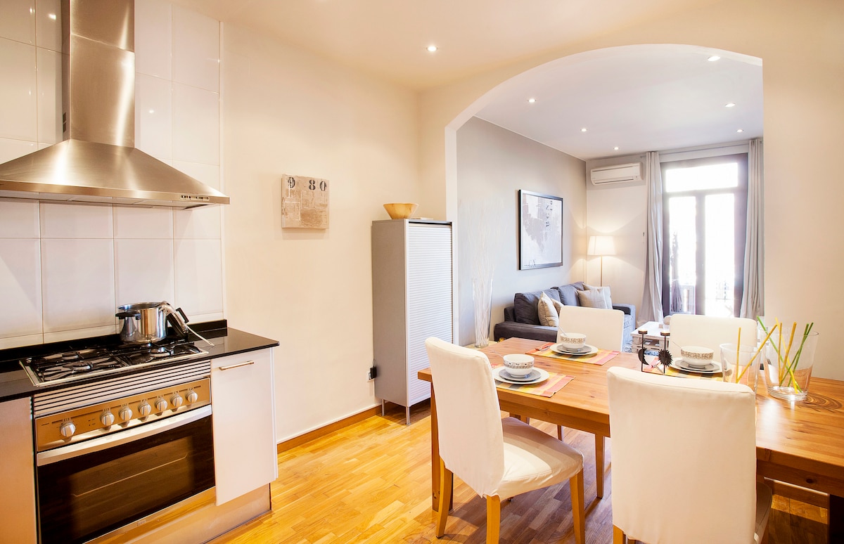 The open kitchen features modern appliances, including a gas stovetop and oven. A wooden dining table is set for four, surrounded by white chairs. A comfortable seating area is visible in the background, illuminated by natural light from a balcony. Neutral tones and light wood flooring create a spacious feel.