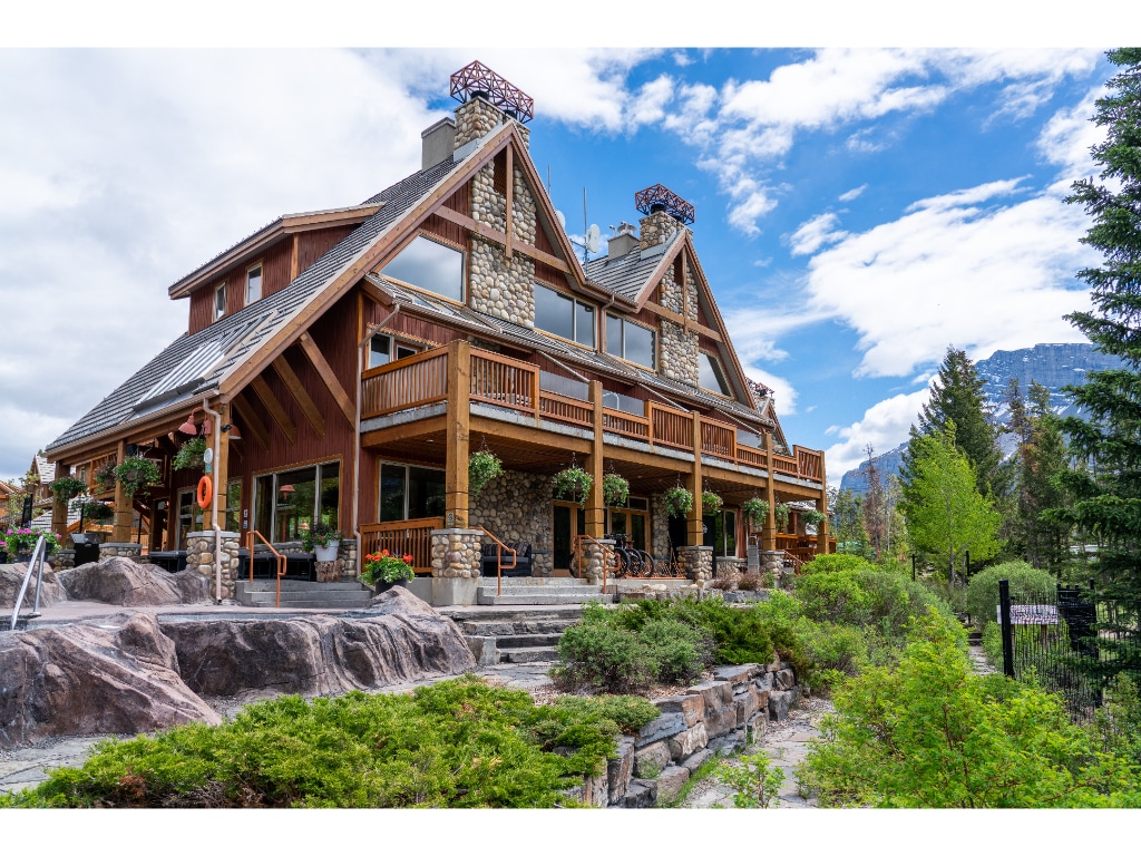 The three-story mountain-style condo is surrounded by lush greenery and rocky landscaping. An expansive deck with wooden railings features potted plants, while large windows provide a view of the outdoors. The iconic architecture is highlighted by stone and wooden elements characteristic of the Rocky Mountain region.