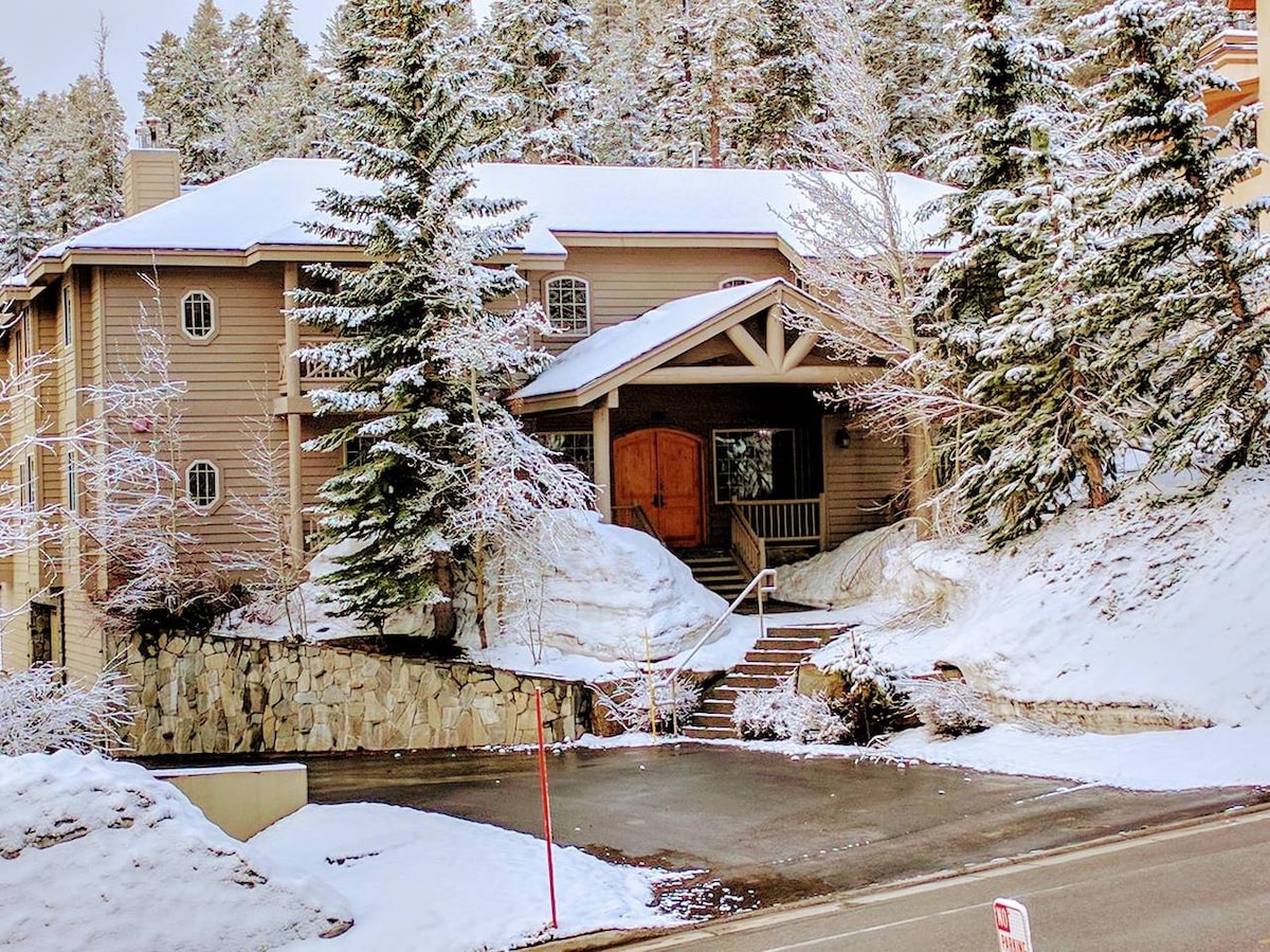 A spacious exterior view of the condo building is framed by snow-covered pine trees. The entrance features a large wooden door and a series of stairs leading up to a covered porch, all surrounded by a blanket of fresh snow.