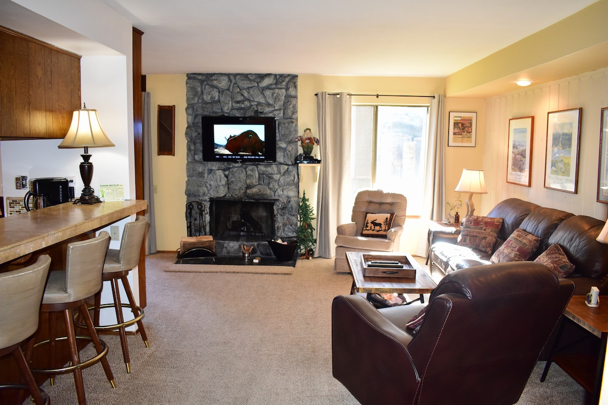 The living room features a stone fireplace and a mounted television above it. A plush sofa and leather chairs provide seating options. Natural light enters through large windows, complemented by framed artwork on the walls. A bar area with tall stools is visible in the background.