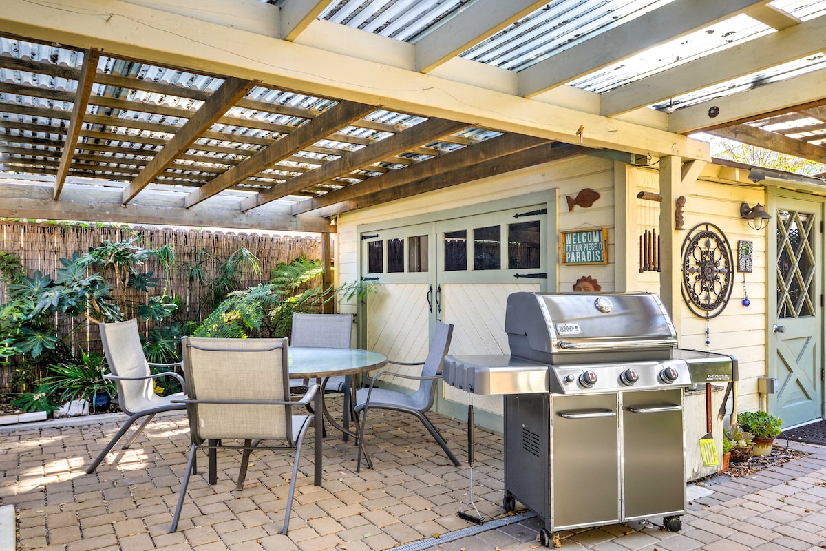 An outdoor dining area is featured under a covered patio, equipped with a glass-topped table and four chairs. A gas grill is positioned nearby, with lush greenery providing a natural backdrop. The space is designed for comfortable al fresco dining and gatherings.