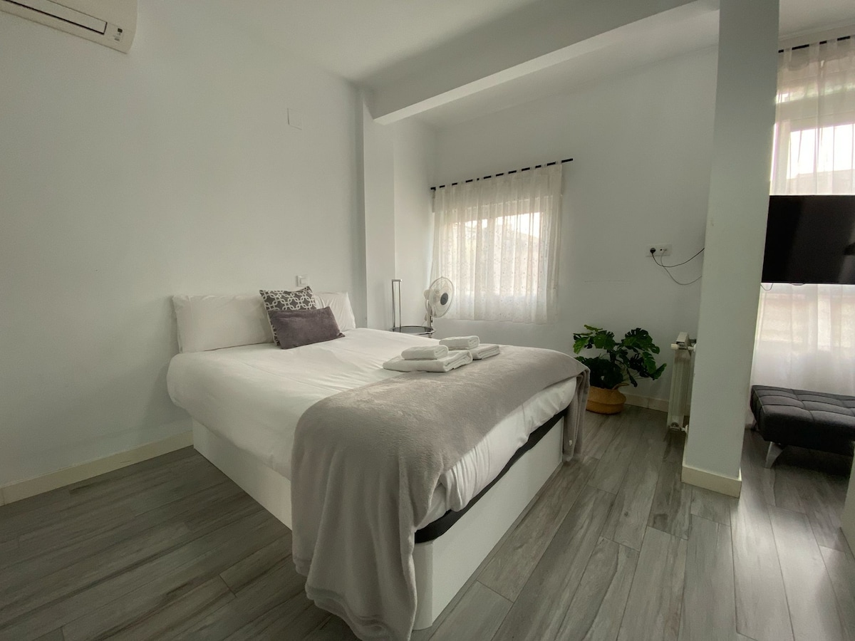 The sleeping area features a spacious bed with neutral-colored linens and decorative cushions. A fan sits on the bedside, and a small plant adds a touch of greenery. Natural light filters through sheer curtains, enhancing the bright, minimalistic ambiance.
