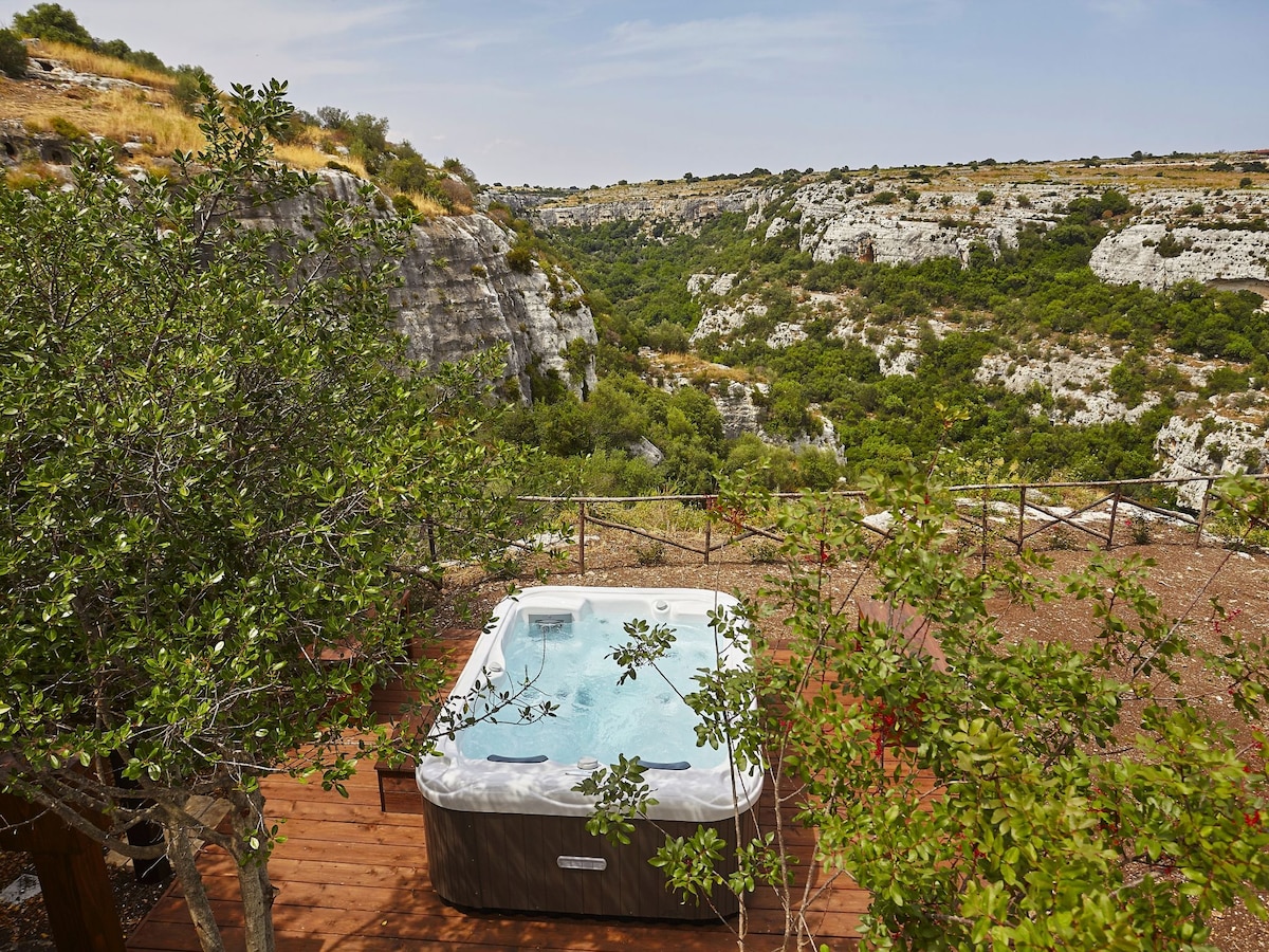 A hot tub is situated on a wooden deck, surrounded by greenery. The scene features a panoramic view of a valley with rocky cliffs, emphasizing the natural landscape. Sunlight illuminates the area, creating a serene outdoor space.