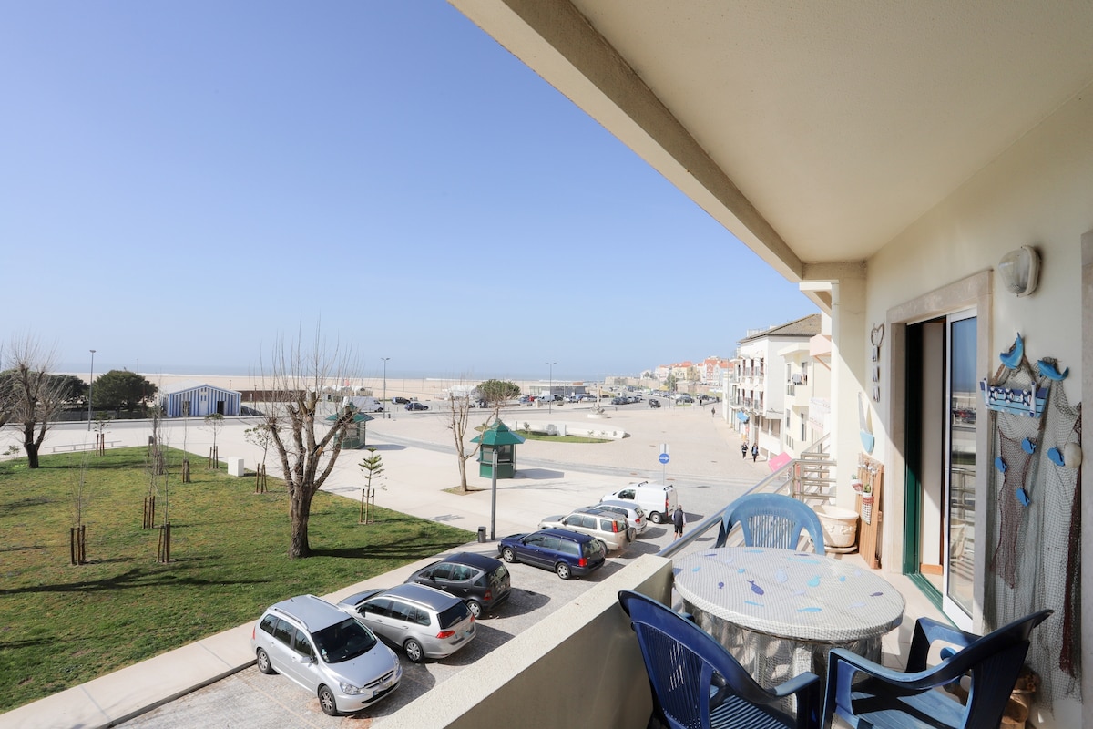 A balcony view highlights a spacious outdoor area, featuring a round table and blue chairs. The landscape includes well-maintained grass and parked cars below, with the beach visible in the distance under a clear blue sky.