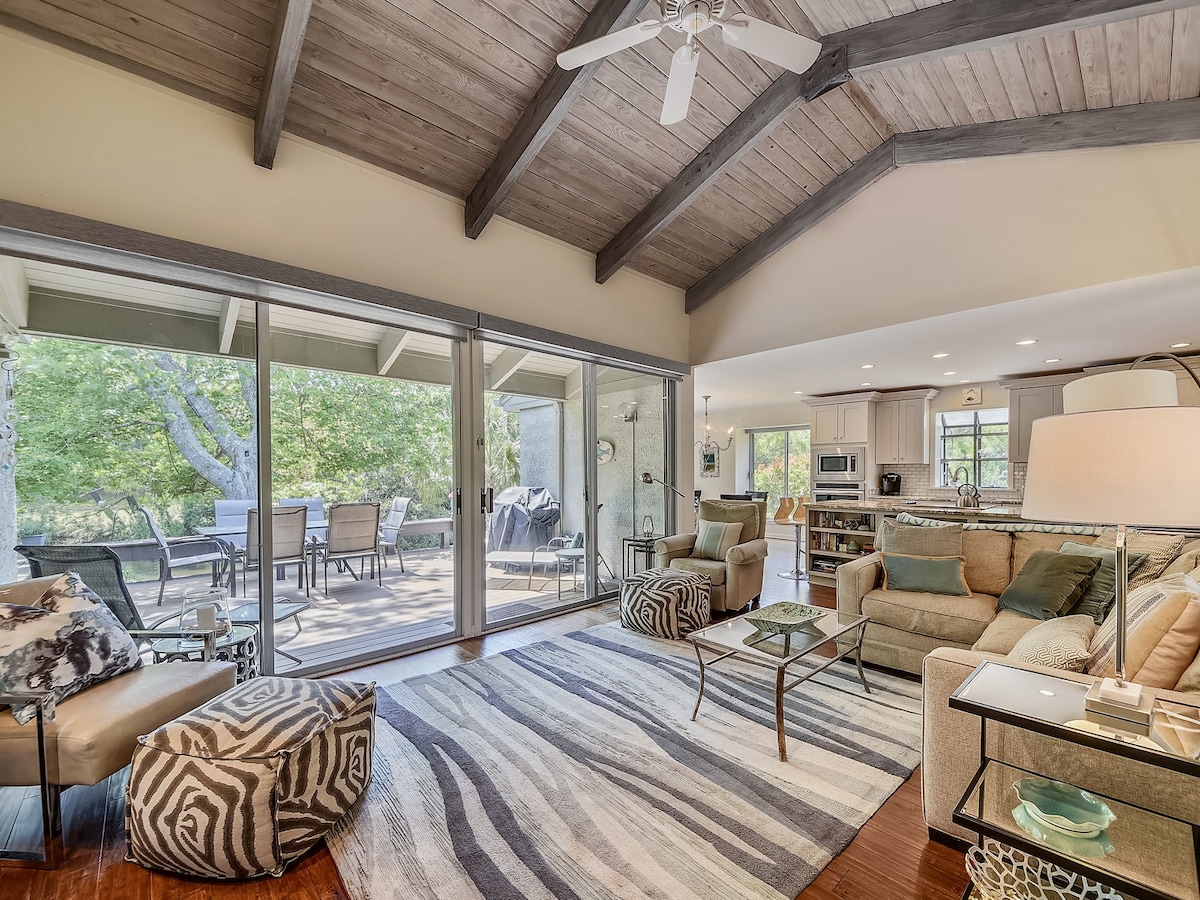A spacious living area features a large couch and two armchairs, arranged around a coffee table. Natural light floods the room through sliding glass doors, revealing a view of the outdoor deck. The room is complemented by a vaulted ceiling and a neutral color palette.