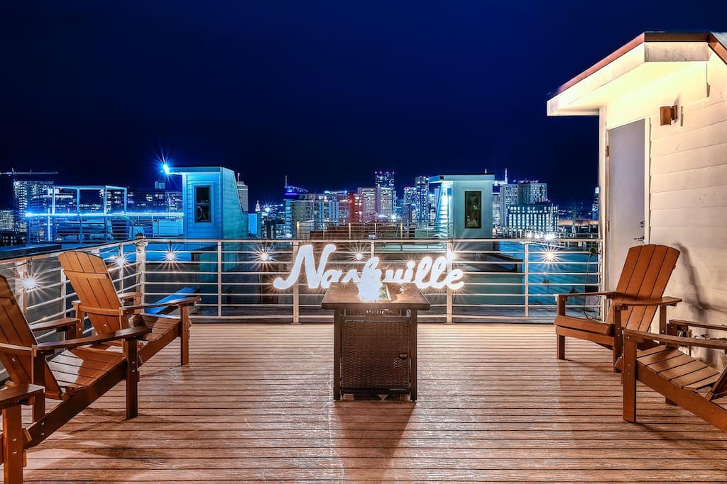 A rooftop deck features two wooden chairs and a small table, with a glowing sign reading 'Nashville' in the background. The skyline of downtown Nashville is illuminated at night, creating a vibrant cityscape against a dark sky.