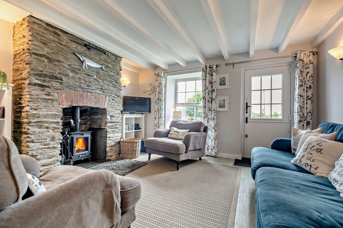 A welcoming living room features exposed beams and a stone fireplace with a woodburner. Comfortable seating options are available, including a brown armchair and a blue couch. Natural light enters through the window, complemented by patterned drapes. A television is mounted on a shelf nearby.
