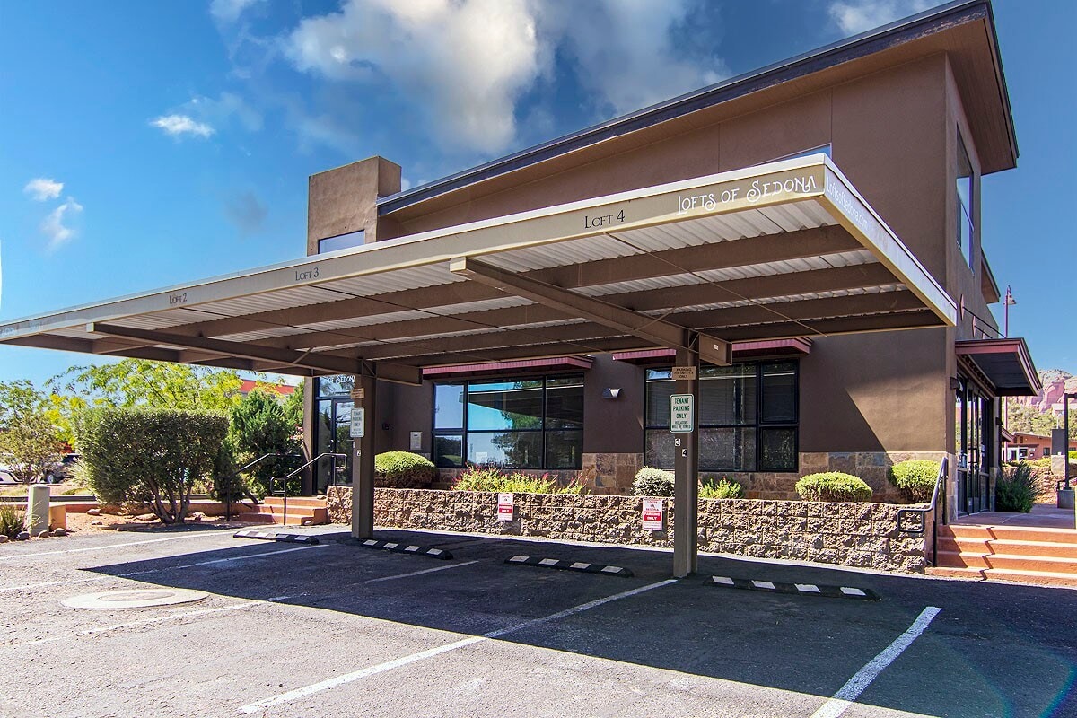 The exterior of the loft building is displayed, featuring a covered parking area. Steps lead up to the entrance, with a landscaped area of low shrubs and trees nearby. Large windows provide a glimpse into the interior space, reflecting natural light.