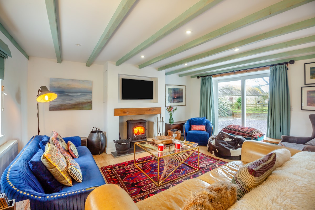 A cozy lounge area is presented with two sofas, one in blue velvet and another in a warm shade, inviting relaxation. A coffee table sits centrally, adorned with candles and decorative items. A woodburner adds warmth, while large glass doors provide views of the surrounding greenery.