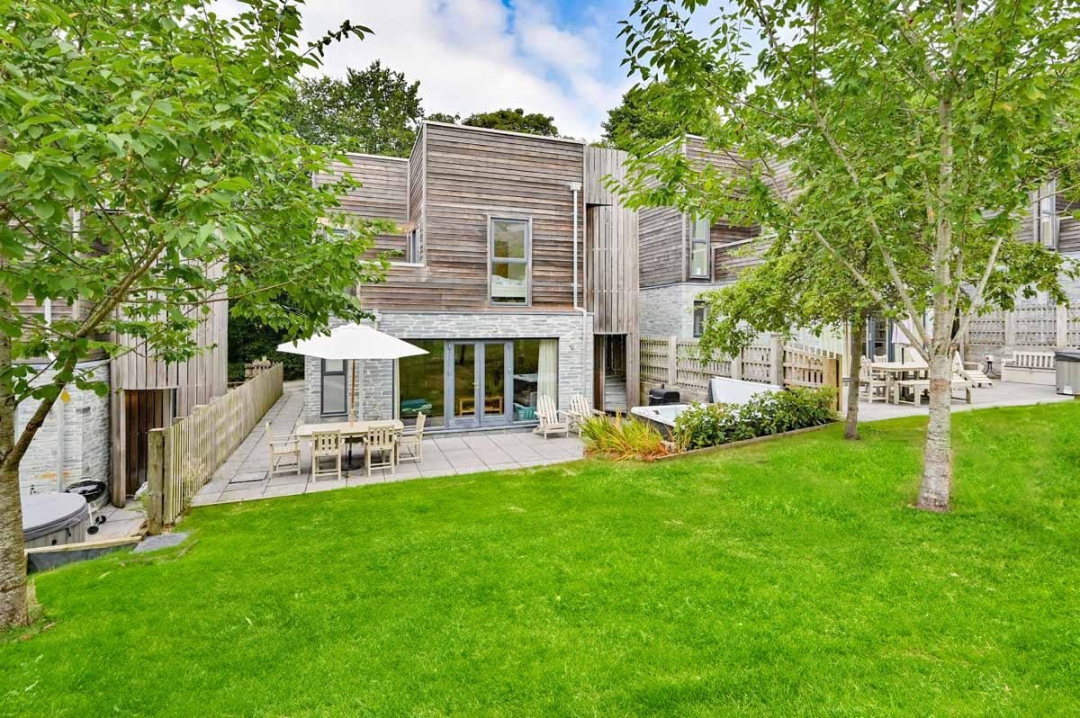 An outdoor view of the property showcases a modern design with natural wood accents. Spacious patios are furnished with dining tables and parasols, surrounded by green lawns and trees. The area offers easy access to a hot tub and plant-filled landscaping, creating an inviting ambiance.