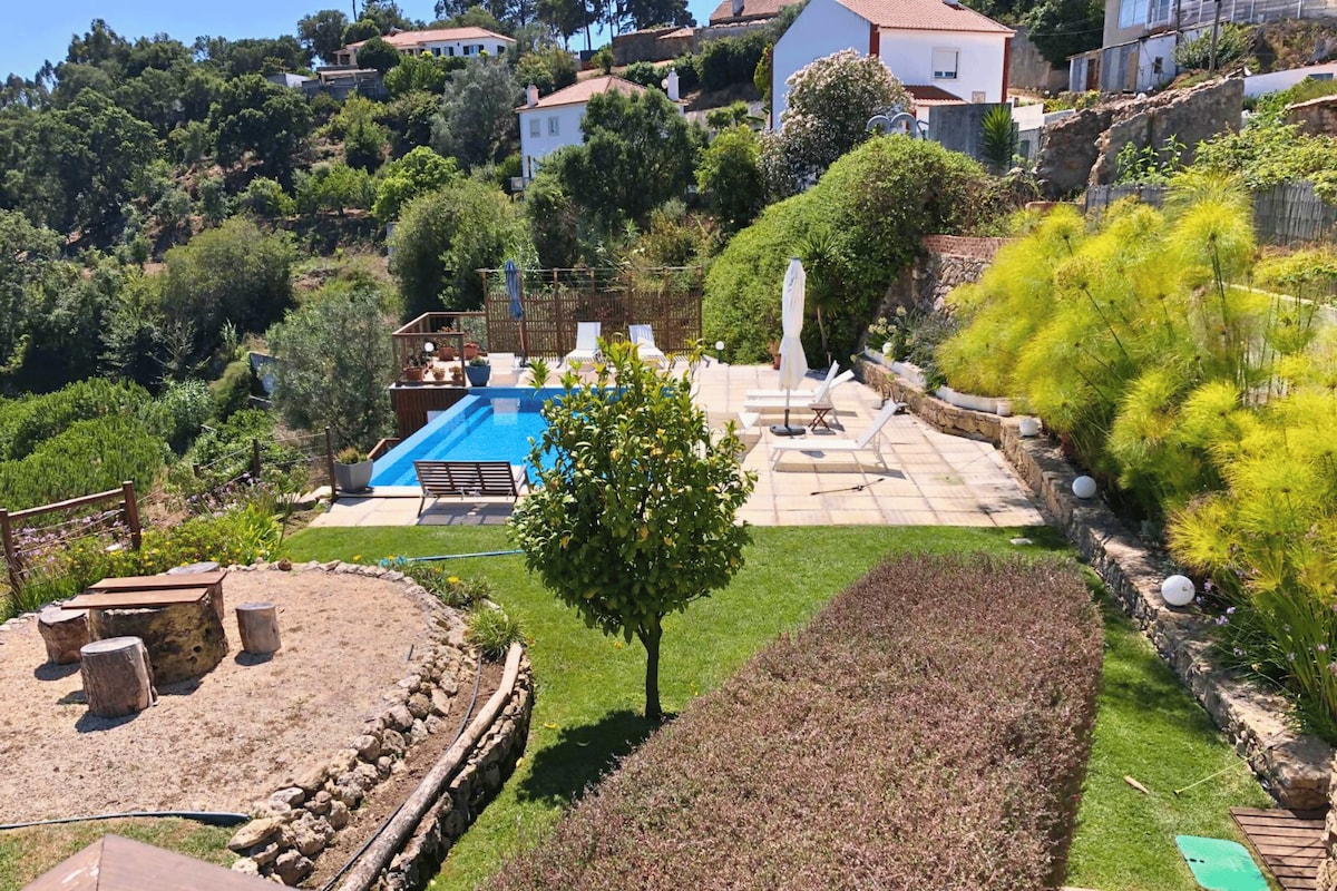 A well-maintained outdoor area features an infinity pool surrounded by sun loungers and umbrellas. Lush greenery and landscaped gardens create a serene atmosphere. A children's play area with natural elements, including a wooden seating area, complements the tranquil setting.