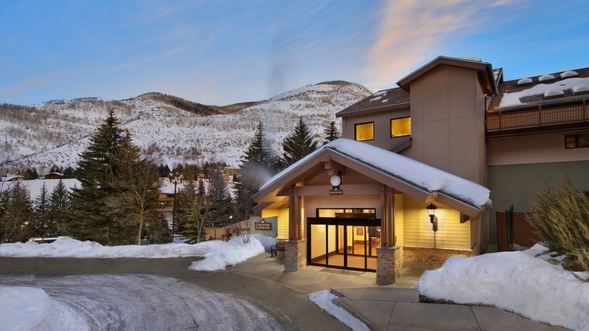 A welcoming entrance is marked by a large doorway framed by stone accents beneath an overhang. Snow-covered mountains rise in the background, hinting at winter activities. Flanking trees add a touch of nature to the inviting facade of the building.