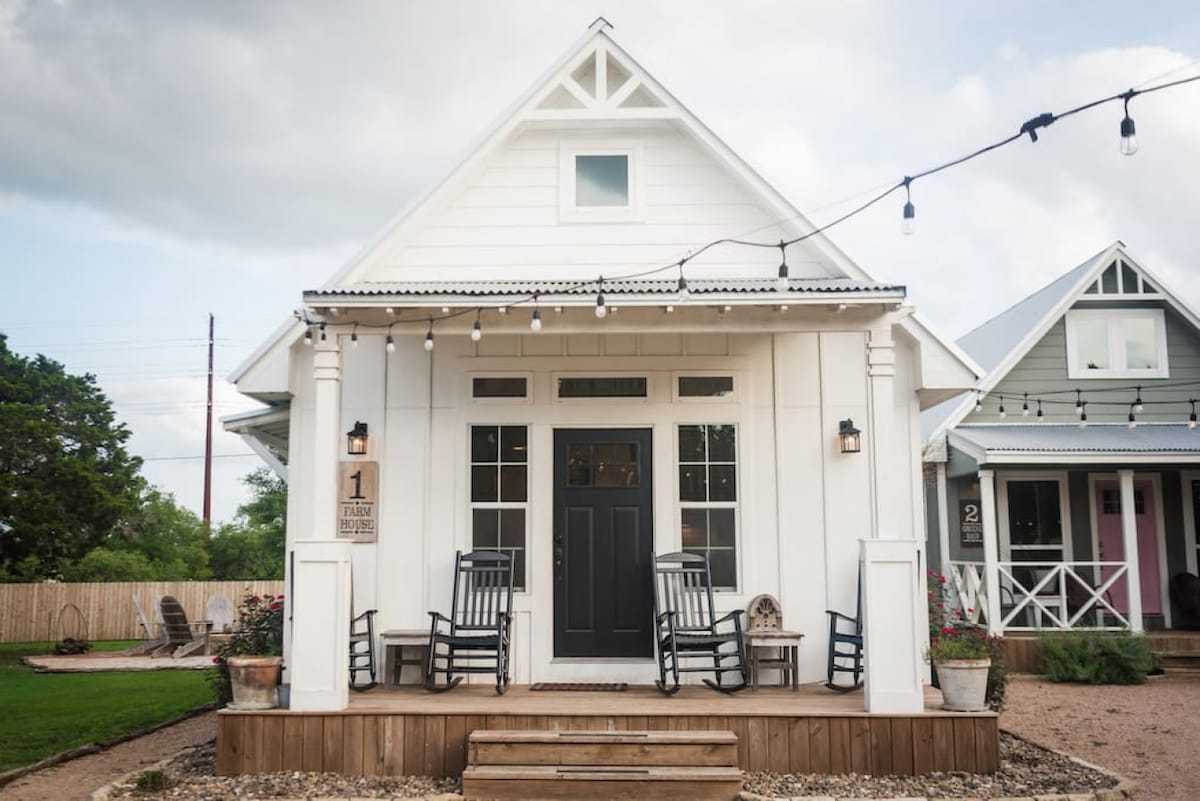 A charming entrance to The Farm House features a dark front door framed by large windows. Rocking chairs are arranged on a wooden porch, inviting relaxation. Decorative lights are strung above, adding a cozy touch to the inviting facade surrounded by a landscaped pathway.