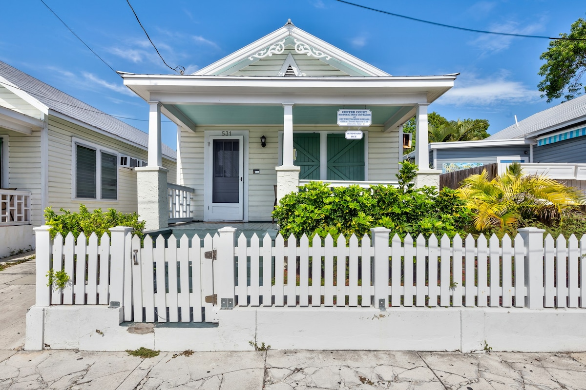 A charming cottage exterior features a white picket fence surrounding a well-maintained garden. The light-colored house is complemented by turquoise accents on the doors and windows, with a covered porch inviting entry. Clear blue skies can be seen overhead.