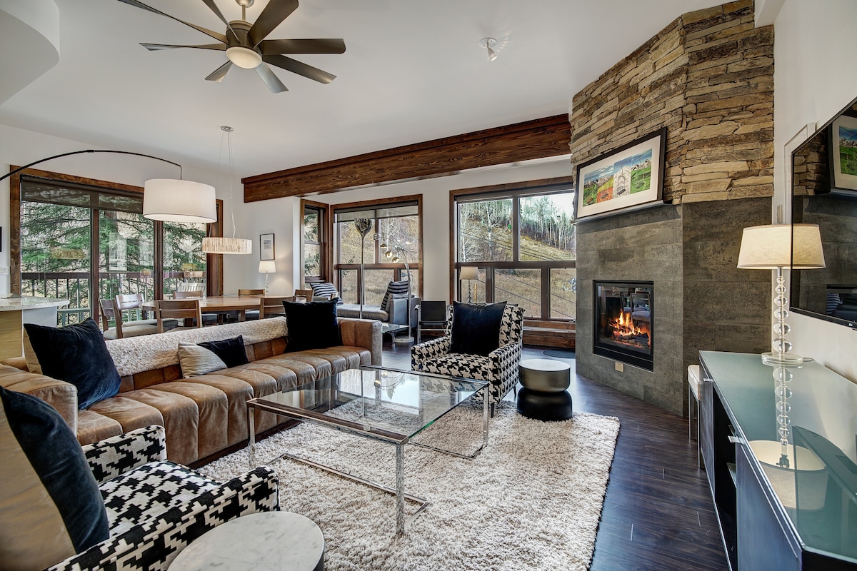 A stylish living area features a combination of comfortable seating, including a leather sofa and patterned chairs. Large windows invite natural light, offering views of the surrounding landscape. A stone fireplace complements the modern decor, and a glass coffee table rests on a plush area rug.