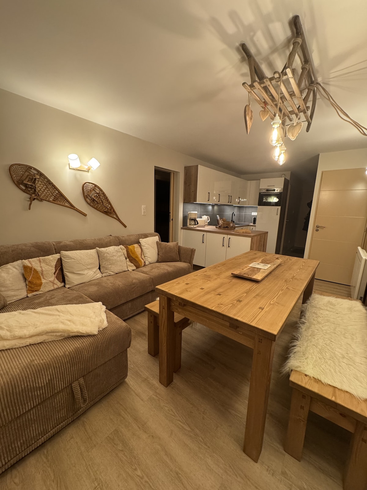 The living area features a comfortable sofa adorned with decorative cushions, facing a rustic wooden dining table. The kitchen is equipped with modern appliances and soft lighting, while ski-themed decor hangs on the wall, adding charm to the space.