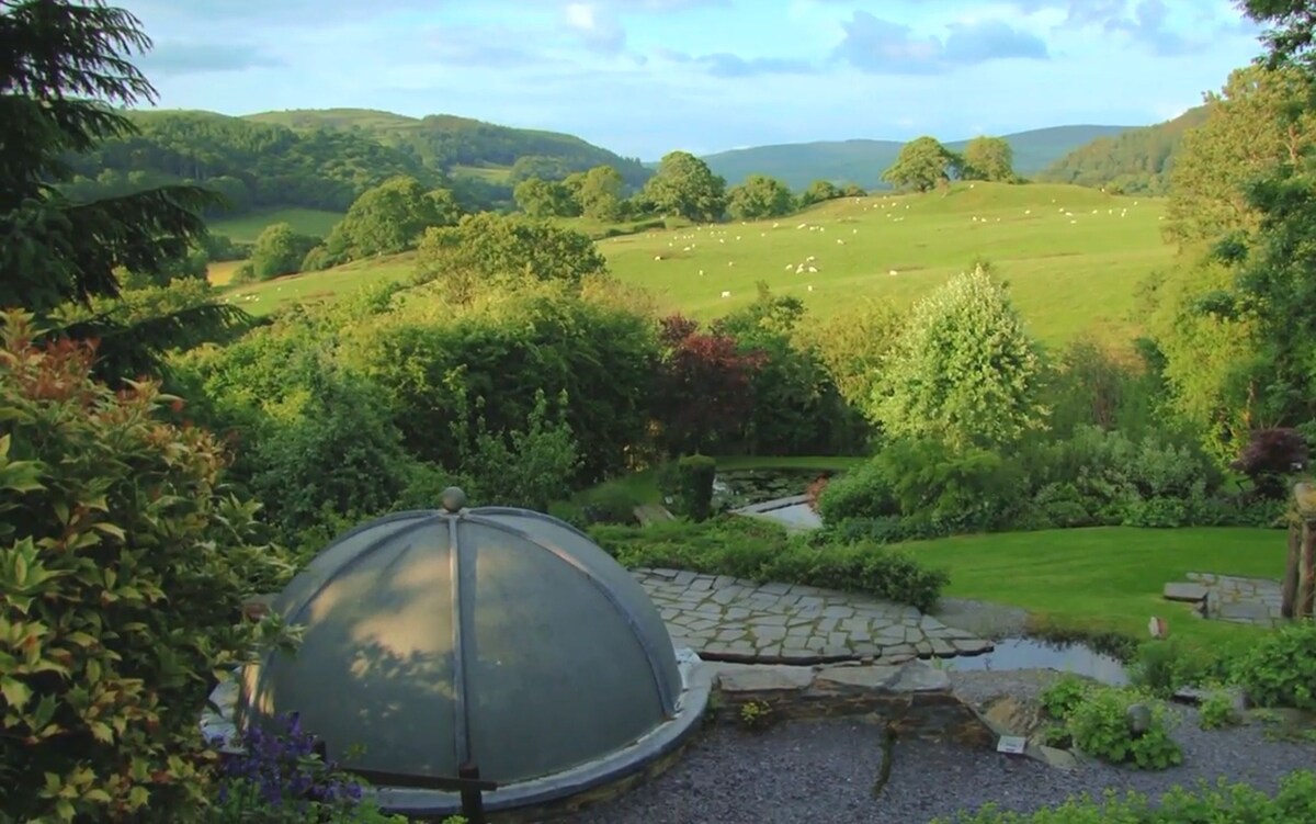 A serene hillside garden is featured, with a decorative dome structure in the foreground. Lush greenery and colorful foliage surround the area, while gentle rolling hills appear in the background, dotted with grazing sheep under a blue sky with scattered clouds.