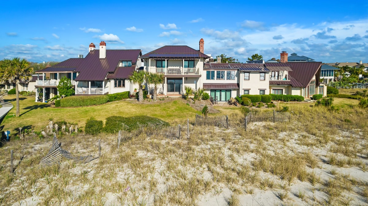 A spacious oceanfront villa is surrounded by a well-maintained landscape, featuring manicured lawns and sandy patches. The structure showcases multiple sections with a combination of stone and stucco finishes, punctuated by chimneys and large windows reflecting the sky's blue hues.