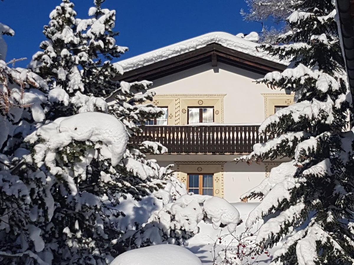 The exterior of a two-family house is visible, partially obscured by snow-covered evergreen trees. The building features a balcony with wooden railings and decorative detailing, set against a clear blue sky. Snow blankets the ground and branches, creating a serene winter landscape.