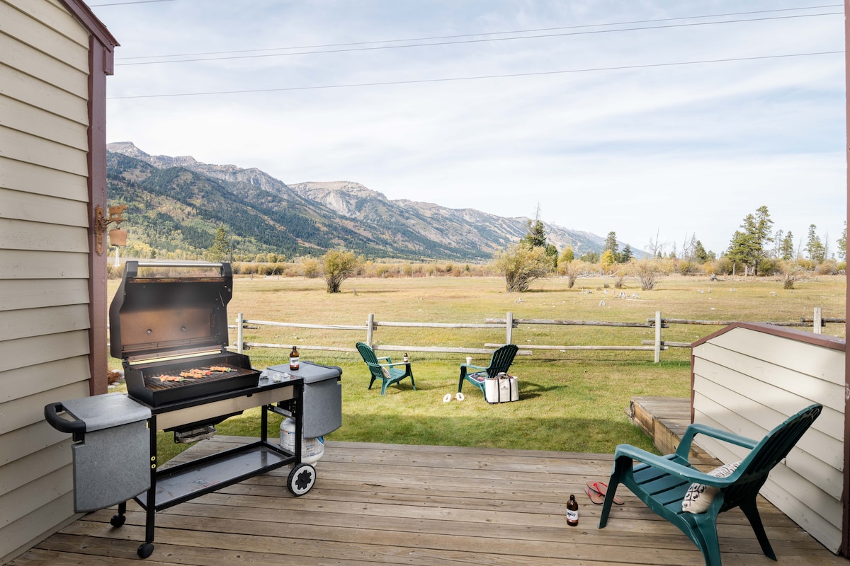 A wooden deck features a charcoal grill, with a cooking surface displaying delicious food. Two green outdoor chairs are positioned nearby, overlooking a spacious lawn and scenic mountain views in the distance. A serene atmosphere is complemented by a few scattered items on the ground.