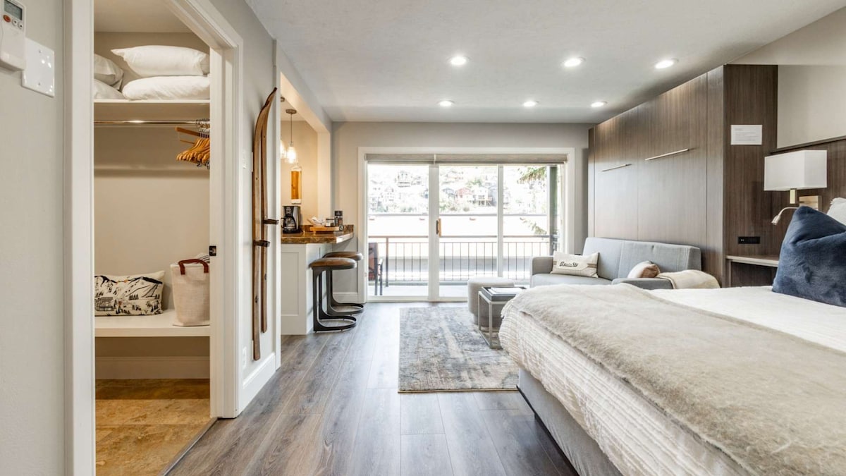 A bright studio space is featured, showcasing a spacious bed and a cozy seating area. Natural light fills the room through sliding glass doors that open to a balcony. A well-organized closet is visible, offering additional storage options. Modern decor adds a touch of elegance.